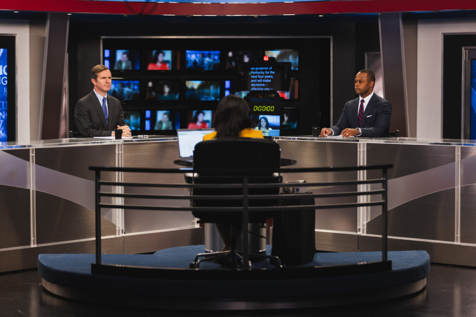 A couple of people sitting at a table in front of some tv screens.